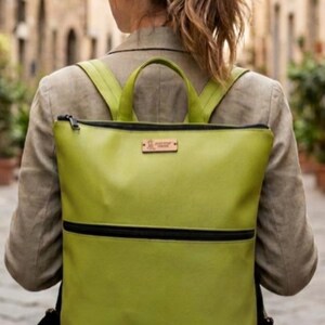 May include: A bright green backpack with black detailing, including a stripe and zip. The bag has two shoulder straps and a top handle. A small wooden tag is attached to the front. The person is wearing a beige blazer and blue jeans.