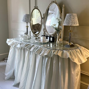 May include: A vanity table featuring a glass top and a floor-length, light blue and white striped fabric skirt with a ruffled edge. The table holds two oval mirrors with ornate gold frames, two lamps with patterned shades, and various glass bottles.