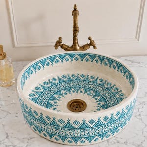 Op de afbeelding: Een ronde, crèmekleurige keramische wastafel met een turquoise geometrisch patroon. De wastafel heeft een messing afvoer en wordt gecombineerd met een messing kraan. Een doorzichtige glazen zeepdispenser met een gouden pomp staat links. De wastafel staat op een marmeren aanrecht.