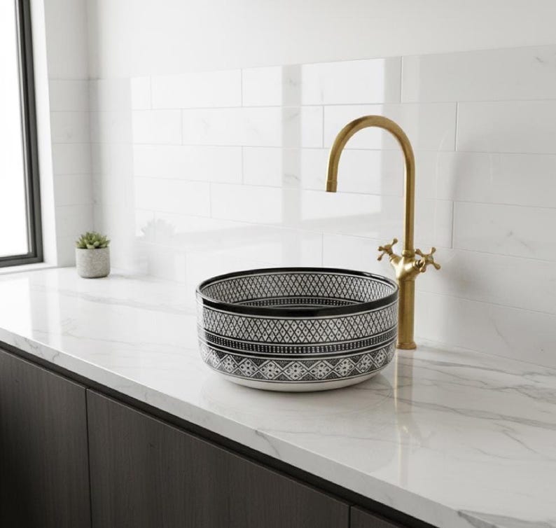 Round Moroccan ceramic vessel sink with hand-painted black-and-white geometric pattern and gold drain on a marble counter.