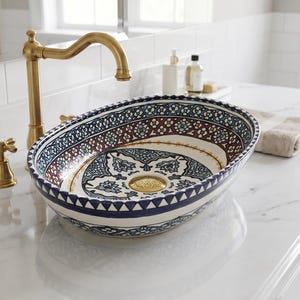 May include: An oval ceramic bathroom sink with a decorative pattern in blue, brown, and white. The sink is set on a white countertop with a gold tap and fixtures. A folded towel rests nearby.