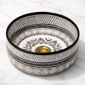 Round Moroccan ceramic vessel sink with hand-painted black-and-white geometric pattern and gold drain on a marble counter.