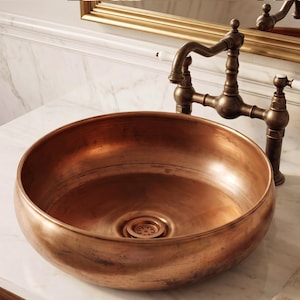 Op de afbeelding: Een ronde koperen waskom met een gepolijste, warme metallic afwerking en een bijpassende centrale afvoer. De kom is gecombineerd met een vintage-stijl donkerbronzen brugkraan met twee witte porseleinen hendels en een gebogen uitloop.