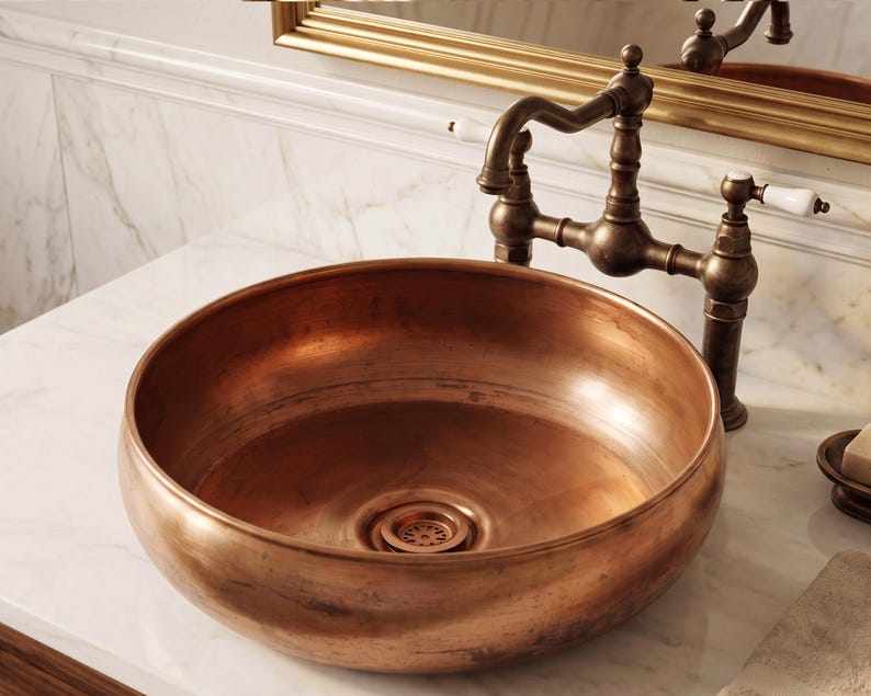 Op de afbeelding: Een ronde koperen waskom met een gepolijste, warme metallic afwerking en een bijpassende centrale afvoer. De kom is gecombineerd met een vintage-stijl donkerbronzen brugkraan met twee witte porseleinen hendels en een gebogen uitloop.