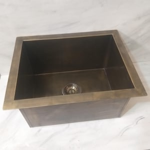 Könnte beinhalten: Ein rechteckiges Küchenbecken in Bronzefarbe mit quadratischem Becken und dekorativem Abfluss. Das Waschbecken hat einen flachen Rand und ein gebürstetes Finish. Das Gesamtdesign ist modern und minimalistisch.