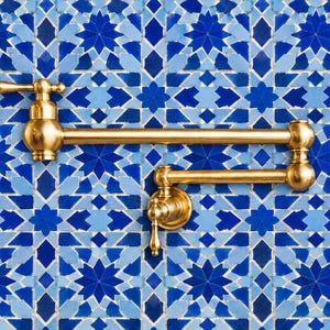 Könnte beinhalten: Ein goldfarbener, wandmontierter Topffüller-Wasserhahn mit einem doppelt gelenkigen, schwenkbaren Arm und zwei separaten Griffen. Die Armatur ist mit einer polierten Metalloberfläche versehen und vor einer blau-weißen Fliesenwand montiert.