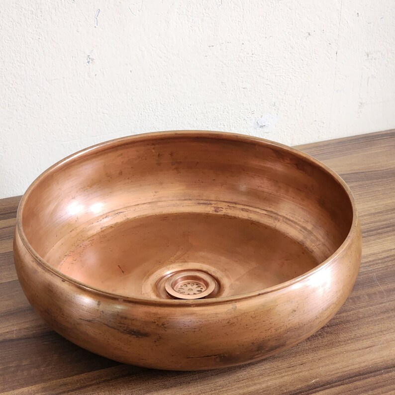 Op de afbeelding: Een ronde koperkleurige waskom met een gladde, gepolijste afwerking en een centrale afvoer voorzien van een decoratief, geperforeerd metalen rooster. De waskom heeft een brede, gebogen rand en een diepe, cirkelvormige binnenkant.