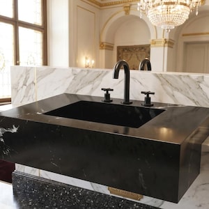 May include: A modern black marble sink with a rectangular basin and a matte black faucet. The sink has two black lever handles and is set against a marble backsplash. The room features a chandelier and ornate architectural details.