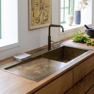 Puede incluir: Un fregadero de cocina rectangular de latón con escurridor integrado, encastrado en una encimera de madera clara. El fregadero cuenta con un grifo de tono bronce oscuro, un paño gris doblado y un pequeño nivel de burbuja.