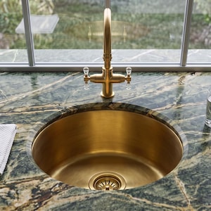 Op de afbeelding: Een ronde messing gootsteen in een donkergroen marmeren aanrechtblad met witte aders. Een hoge messing kraan met heldere kristallen handgrepen staat achter de gootsteen. Een gevouwen witte textiel doek en een glas water staan op het aanrecht.