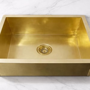 Op de afbeelding: Een rechthoekige, goudkleurige gootsteen met een afvoer. De gootsteen heeft een geborstelde afwerking en staat op een wit marmeren aanrecht. De afvoer is cirkelvormig met een decoratief patroon.