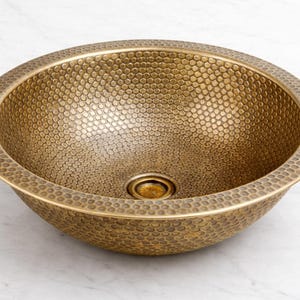May include: A round, bowl-shaped sink featuring a textured, hammered finish across its entire surface. The basin is crafted from a metallic material with a warm, antique brass tone and includes a central drain opening at the bottom of the interior.