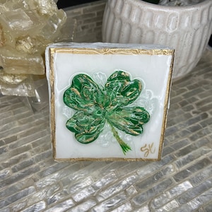 May include: A four-leaf clover with gold accents is painted on a white square canvas with a gold frame. The canvas is sitting on a white brick surface.