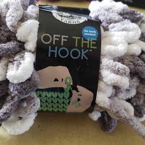 May include: A skein of chunky, looped yarn in a variegated mix of white, light gray, and dark gray. A black paper label wraps around the center, featuring the text OFF THE HOOK, a graphic illustration of hands knitting, and a blue circle stating No tools needed.
