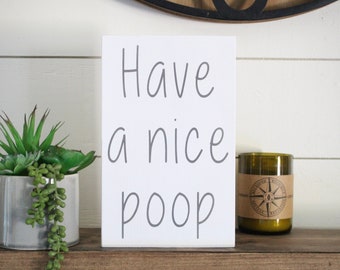May include: White wooden sign with the text "Have a nice poop" in gray cursive lettering. The sign is sitting on a wooden shelf with a small potted succulent plant and a green glass candle to the right.