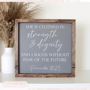 May include: A gray wooden sign with a white inscription that reads "She is clothed in strength & dignity and laughs without fear of the future. Proverbs 31:25".