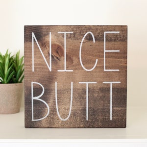 May include: A wooden sign with the text "NICE BUTT" painted in white on a brown background.