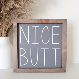 May include: A wooden sign with a gray background and white text that reads "NICE BUTT".