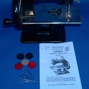 May include: A black Singer 20-10 sewing machine with a silver metal bedplate and a black plastic base. The machine is accompanied by a set of needles, bobbins, and instructions for use. The instructions include the text "INSTRUCTIONS FOR USING SINGER SEWING MACHINE 20-10" and "TO ALL WHOM IT MAY CONCERN... THE SINGER MANUFACTURING COMPANY."