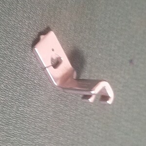 May include: A silver metal sewing machine foot with a curved shape and a small hole in the top.
