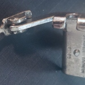 May include: A silver sewing machine foot with a lever and the text "SINGER TAIWAN" printed on the side.