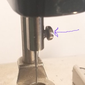May include: Close-up of a sewing machine needle and needle clamp. The needle is threaded and the clamp is secured with a screw.