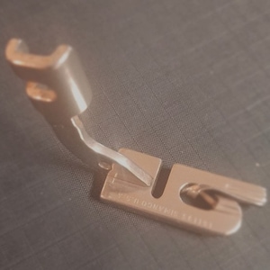 May include: A silver sewing machine foot with a curved metal arm and a flat base. The base has the text "SINGER 1/4 INCH" printed on it.