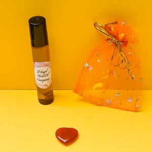 Può includere: Un piccolo flacone di vetro ambrato con un applicatore a rullo nero e un'etichetta bianca con la scritta "Angel Mood Oil Energising". Il flacone è su una superficie gialla accanto a una piccola borsa di rete arancione con un nastro dorato e una pietra rossa a forma di cuore.