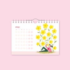 Pode incluir: Um calendário para o mês de maio com fundo rosa. O calendário apresenta uma ilustração de uma pessoa relaxando entre grandes flores amarelas. A palavra "May" é impressa no topo do calendário.