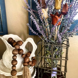 May include: A bouquet of wooden tulips in shades of brown, beige, and red, arranged in a wire basket with lavender sprigs. The bouquet is sitting on a table next to a white pitcher with a brown wooden bead garland.