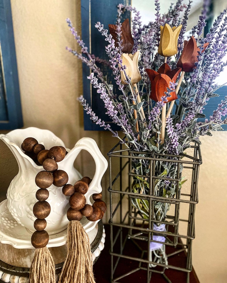 May include: A bouquet of wooden tulips in shades of brown, beige, and red, arranged in a wire basket with lavender sprigs. The bouquet is sitting on a table next to a white pitcher with a brown wooden bead garland.