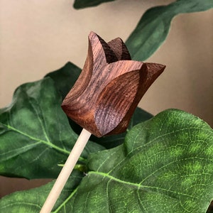 May include: A wooden tulip flower with a brown wood grain finish. The flower is attached to a thin wooden stick.