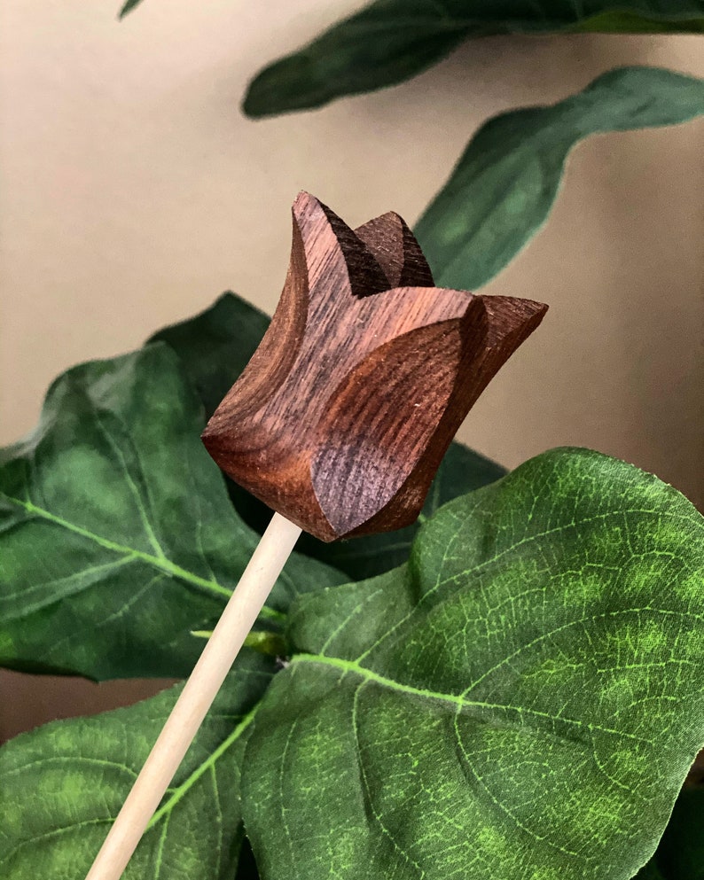 May include: A wooden tulip flower with a brown wood grain finish. The flower is attached to a thin wooden stick.