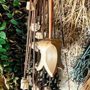 May include: A wooden flower-shaped ornament hanging from dried flowers and greenery. The ornament is light brown and has a pointed tip.