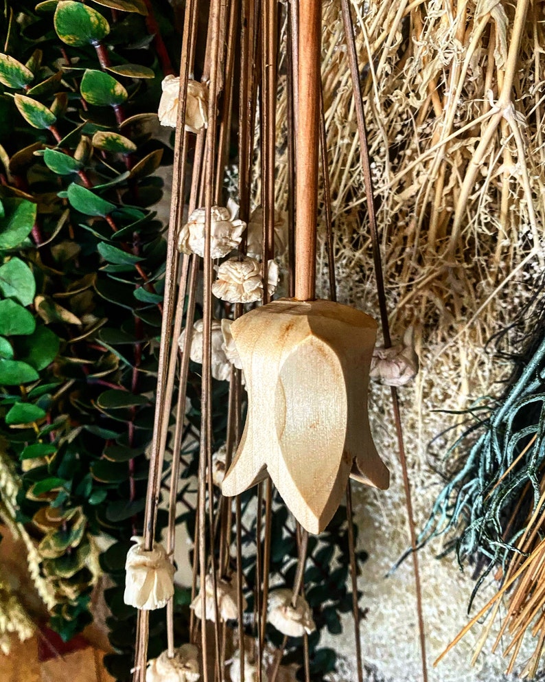 May include: A wooden flower-shaped ornament hanging from dried flowers and greenery. The ornament is light brown and has a pointed tip.