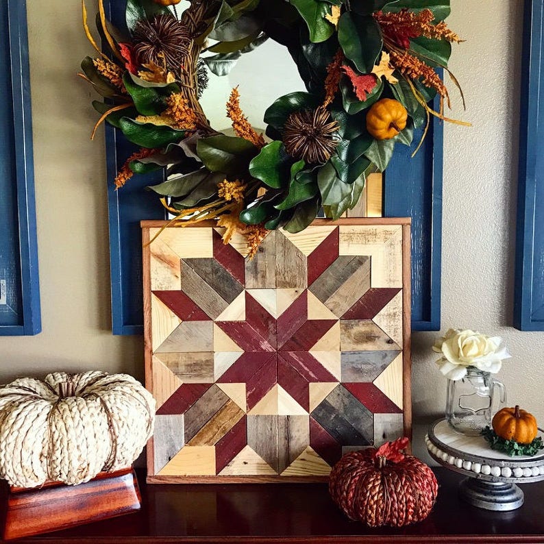 Puede incluir: Un tapiz de pared de madera con un patr&oacute;n geom&eacute;trico rojo, marr&oacute;n y blanco. El tapiz est&aacute; enmarcado en madera y est&aacute; rodeado de decoraci&oacute;n de oto&ntilde;o, incluyendo una corona con calabazas y una calabaza blanca.