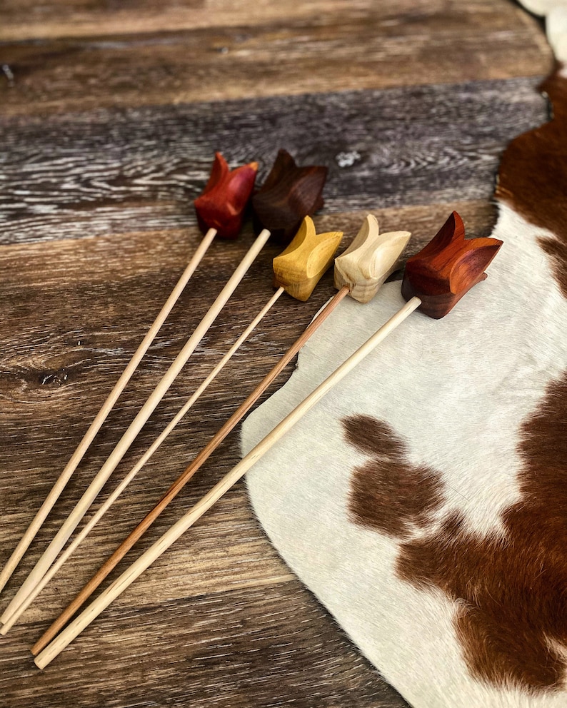 May include: Five wooden tulip-shaped flower picks in various colors, including red, brown, yellow, and natural wood. The picks are arranged on a wooden surface with a cowhide rug in the background.