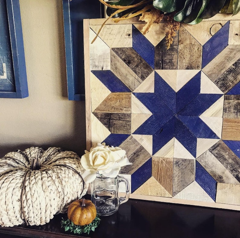 Puede incluir: Una pieza de arte de pared de madera con un dise&ntilde;o de estrella azul y blanca. La estrella est&aacute; hecha de cuadrados de madera m&aacute;s peque&ntilde;os, cada uno pintado de un tono diferente de azul. El fondo es de madera marr&oacute;n claro.