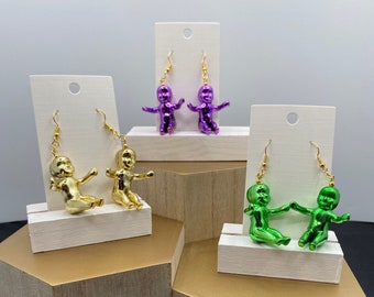May include: Three pairs of earrings featuring a baby doll design. The earrings are displayed on white stands with a gold, green, and purple finish.