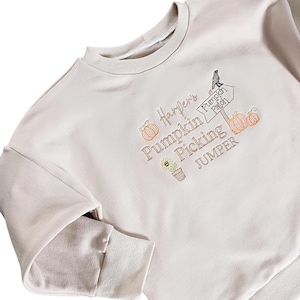May include: A beige sweatshirt with an embroidered design that says "Harper's Pumpkin Picking Jumper" with a pumpkin, sunflower, and a sign that says "Pumpkin Patch".