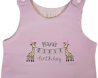 May include: A light purple sleeveless onesie with a personalized embroidered design featuring two giraffes wearing party hats and a banner that reads "Maya's First Birthday".