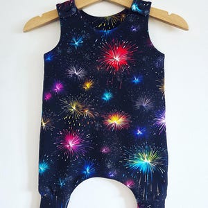 May include: A navy blue baby romper with a vibrant fireworks pattern in shades of red, pink, yellow, blue, and green. The sleeveless romper has a round neckline, snap closures on the shoulders, and elastic cuffs at the ankles.