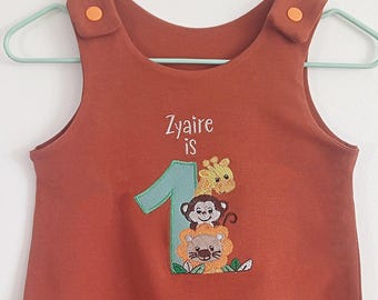 May include: A rust-colored sleeveless onesie with a green number one, a giraffe, a monkey, and a lion embroidered on the front. The text "Zyaire is" is embroidered above the number one.