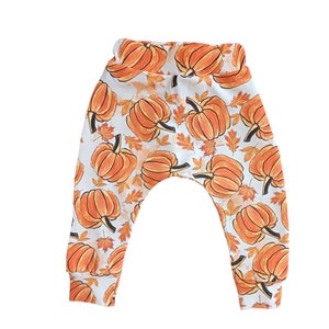 Peut inclure: Pantalon de bébé avec un imprimé intégral de citrouilles oranges et de feuilles d'automne sur fond blanc. Le pantalon a une large ceinture et des poignets ajustés. Parfait pour l'automne.