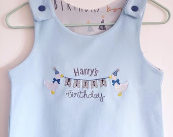 May include: Light blue sleeveless onesie with a white and gray embroidered banner that says "Harry's First Birthday" with two white ducks wearing party hats on either side of the banner.