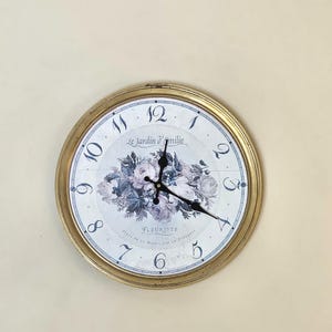 May include: A round wall clock with a gold-colored frame. The clock face features a floral design in shades of purple and white, with black clock hands and Roman numerals. The text "Le Jardin d'Emilie" and "Fleuriste" are visible.