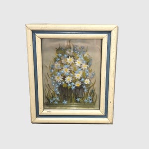 May include: Framed floral painting featuring a bouquet of white daisies and blue forget-me-nots in a dark gray pot. The artwork is set against a light background and framed in a white and blue wooden frame, creating a vintage aesthetic.
