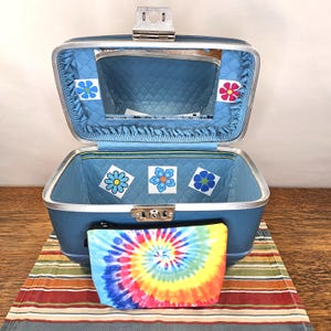 May include: Open blue vintage suitcase with a silver clasp and a mirror inside. The interior is lined with blue fabric and decorated with flower stickers. A tie-dye cosmetic bag rests in front of the suitcase on a striped mat.
