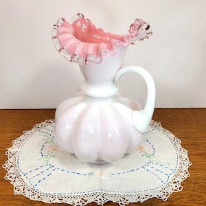 May include: A white milk glass pitcher with a pink ruffled top and a white handle. The pitcher is sitting on a white doily with blue embroidery.