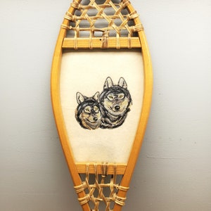 May include: A wooden snowshoe with a woven design and a white fabric panel featuring an embroidered image of two wolves' heads.